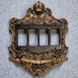 May include: A four-gang light switch wall plate featuring an ornate gold-painted frame with intricate scrollwork and a crown motif at the top. The central rectangular section is dark brown with four vertical openings and visible screw holes.