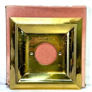May include: A square, polished brass light switch wall plate featuring a beveled edge with a decorative ribbed border and a central circular opening. The plate is positioned against a faded pink cardboard box with visible text on the top edge.