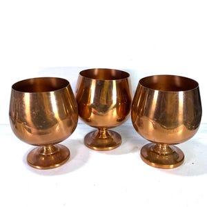 May include: Three copper-colored brandy snifters with a rounded bowl and a short stem. The glasses have a shiny, reflective surface and are arranged on a white background. The snifters are a classic design for serving brandy or cognac.