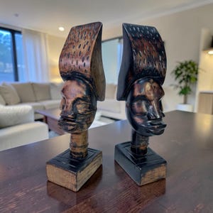 Puede incluir: Dos esculturas de madera talladas a mano de cabezas humanas, cada una con un tocado alargado y texturizado en tonos marrón oscuro y negro. Las figuras están montadas sobre bases rectangulares de madera, mostrando la veta natural de la madera.