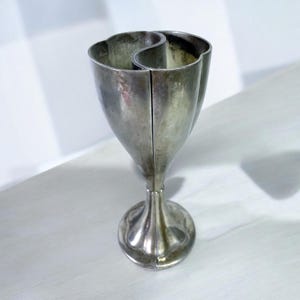 May include: A silver-colored, tulip-shaped vase with a unique, divided design. The vase has a flared top with two curved openings, a slender stem, and a round base. The metallic finish gives it a vintage or antique appearance.