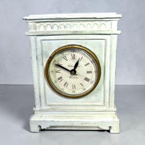 May include: A rectangular mantel clock with a distressed off-white finish and a gold-toned circular bezel. The clock face features black Roman numerals, Arabic minute markers, and the text Richard and Winchester in black script lettering.