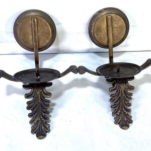 May include: Two antique brass wall sconces. Each sconce features a circular backplate, a curved arm holding a candle cup, and a decorative leaf design. The sconces are made of brass and have a vintage appearance.