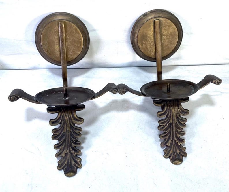 May include: Two antique brass wall sconces. Each sconce features a circular backplate, a curved arm holding a candle cup, and a decorative leaf design. The sconces are made of brass and have a vintage appearance.