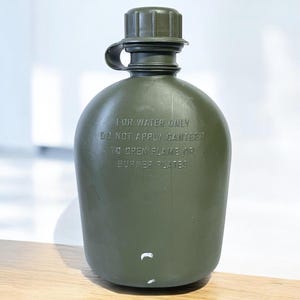 Può includere: Borraccia verde oliva con tappo a vite e coperchio attaccato. La borraccia presenta il testo "FOR WATER ONLY DO NOT APPLY CANTEEN TO OPEN FLAME OR BURNER PLATES" in rilievo sul davanti. La borraccia è realizzata in materiale plastico resistente.