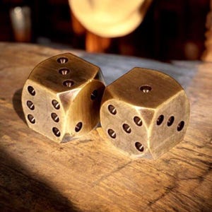 May include: Two antique brass dice with rounded edges, each displaying a different number of recessed dots. The dice are set on a wooden surface, with warm lighting highlighting their metallic finish. A classic game accessory.