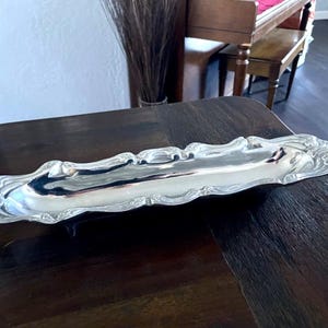 Op de afbeelding: Een zilverkleurige, langwerpige serveerschaal met sierlijke, krullende randen. De schaal is zeer reflecterend, met een glad oppervlak. De decoratieve schaal staat op een donkere houten tafel, wat wijst op een vintage of antieke uitstraling.