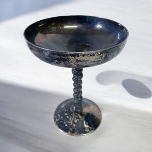 Könnte beinhalten: Eine silberfarbene Champagnerschale mit einer breiten, flachen Schale und einem gedrehten Stiel. Der Sockel ist rund und das Metall hat ein angelaufenes Aussehen, was auf Alter hindeutet. Die Schale ist zum Servieren von Sekt oder Cocktails bestimmt.