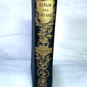 May include: A black hardcover book spine featuring gold embossed lettering and decorative floral patterns. The text reads BERLIN AND SANS-SOUCI, MUHLBACH, and APPLETONS. The design includes a shield-shaped frame at the top and ornate scrollwork.