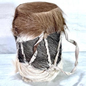 Könnte beinhalten: Kleine, zylindrische Trommel mit braunem Fell oben und einem Körper, der mit schwarz-weißem Fell umwickelt ist. Die Trommel ist mit weißen Lederriemen befestigt und hat einen gedrehten Ledergriff.
