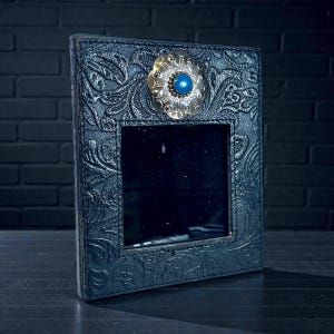 May include: A black leather picture frame with an embossed floral pattern. The frame has a silver-toned, flower-shaped embellishment with a blue centre stone. The frame is rectangular and stands upright on a dark surface.