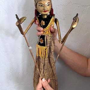 May include: A wooden puppet with a painted face, wearing a black top with gold accents and a patterned skirt. The puppet has jointed arms and is held by wooden rods. The head is adorned with a crown.