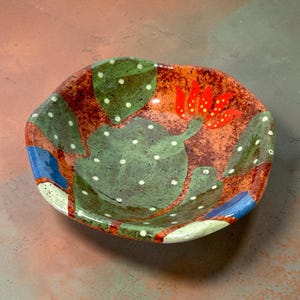 May include: A shallow, hand-painted ceramic bowl with an irregular, organic shape. The interior features a speckled terracotta-colored base decorated with green cactus pads adorned with white dots and a vibrant red flower with yellow accents.