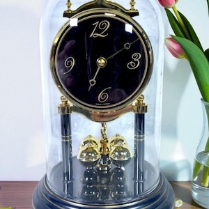 May include: A black and gold clock with a glass dome. The clock face has gold numerals and hands. The base is dark blue with gold trim. Two pink tulips are in a vase to the right of the clock.