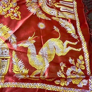 May include: A red textile with intricate gold and white embroidery. The design features a leaping deer, floral patterns, and ornate borders. The fabric has a silky sheen and a fringed edge, suggesting a decorative scarf or shawl.