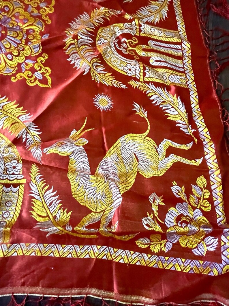 May include: A red textile with intricate gold and white embroidery. The design features a leaping deer, floral patterns, and ornate borders. The fabric has a silky sheen and a fringed edge, suggesting a decorative scarf or shawl.