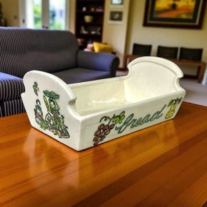 Può includere: Cestino per il pane in ceramica color crema, di forma rettangolare con estremità curve. Il fronte presenta la scritta "Bread" in verde, con illustrazioni di uva e una pera. I lati presentano immagini colorate di persone.