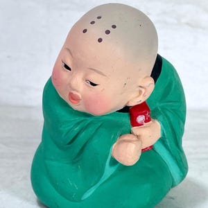 May include: A ceramic figurine of a seated monk in a green robe, holding a red object. The monk has a bald head with dark spots and rosy cheeks. The figurine is a traditional Asian decorative item.