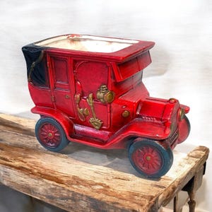 May include: A vintage-style red car-shaped planter with black accents. The planter features gold-colored decorative elements and dark blue wheels. The car is sitting on a wooden surface. This decorative planter is ideal for adding a touch of nostalgia to any space.