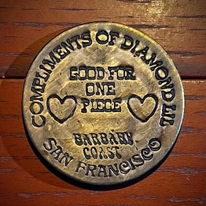May include: A vintage brass coin with the text "COMPLIMENTS OF DIAMOND LIL" and "GOOD FOR ONE PIECE". The coin also features two heart symbols and the text "BARBARY COAST SAN FRANCISCO".