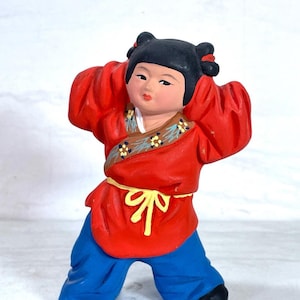May include: A ceramic figurine of a child in a traditional outfit. The figure wears a red top with a yellow belt and blue pants. The child has black hair styled in two buns and is holding their hands up near their head.