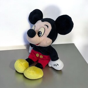 Puede incluir: Un peluche de Mickey Mouse, con orejas negras, cuerpo negro, pantalones cortos rojos con botones amarillos y pies amarillos. El juguete tiene una cara color crema y guantes blancos. El personaje ic&oacute;nico est&aacute; sentado sobre una superficie reflectante.