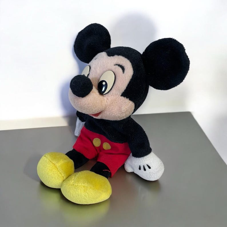 Puede incluir: Un peluche de Mickey Mouse, con orejas negras, cuerpo negro, pantalones cortos rojos con botones amarillos y pies amarillos. El juguete tiene una cara color crema y guantes blancos. El personaje ic&oacute;nico est&aacute; sentado sobre una superficie reflectante.