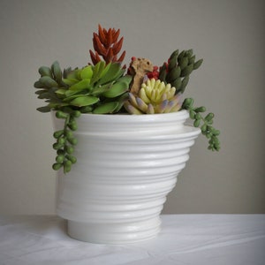 May include: A white ceramic planter with a wavy, textured design, filled with a variety of green, red, and yellow succulents.