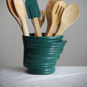 May include: A teal ceramic utensil holder with a unique spiral design, holding a variety of wooden kitchen utensils, including a spatula, spoons, and a wooden spoon with holes.