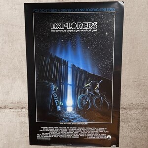 May include: A vertical movie poster for Explorers featuring a night sky with stars above a wooden fence. A bright blue light shines through a gap in the fence. A bicycle and a skateboard lean against the fence. The text reads Explorers and The adventure begins in your own backyard.