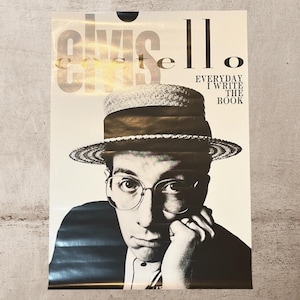 May include: Poster featuring a black and white portrait of Elvis Costello wearing glasses and a hat. The poster has the text "Elvis Costello" at the top and "Everyday I Write The Book" on the right side.