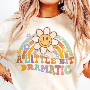 May include: A white t-shirt with a rainbow and a smiley face flower design. The text "A Little Bit Dramatic" is printed in colorful letters on the shirt.