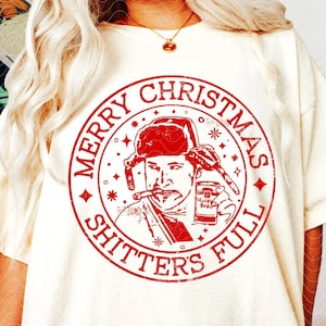 以下が含まれることがあります： クリーム色のTシャツ。赤色のグラフィックは、冬の帽子をかぶり、葉巻を吸い、ビールを持っている男性が描かれています。「MERRY CHRISTMAS SHITTERS FULL」の文字が円形にデザインされています。