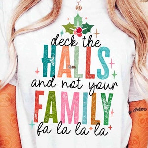 Puede incluir: Una camiseta blanca con la frase "deck the HALLS and not your FAMILY fa la la la" en un diseño festivo y multicolor. El diseño incluye acebo y bayas, ideal para Navidad. La camiseta es una descarga digital.