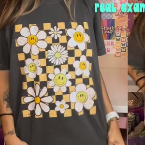 K&ouml;nnte beinhalten: Ein graues T-Shirt mit einem karierten Muster und G&auml;nsebl&uuml;mchen mit Smiley-Gesichtern. Der Text "kinda panda studio" und "PANDACREATIVE.ORG" ist auf dem T-Shirt aufgedruckt. Der Text "real example" ist in gr&ouml;&szlig;erer Schrift gedruckt.