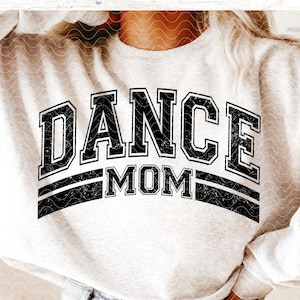 May include: A white sweatshirt with black text that reads "DANCE MOM" in a distressed, varsity-style font. The text is layered over two black stripes.