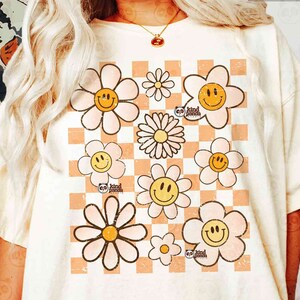 May include: A white t-shirt with a checkered pattern in peach and white. The pattern is overlaid with a design of pink and white daisies with yellow smiley faces. The text "be kind" is written in brown on a small circle in the center of the design.