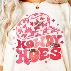 May include: A white t-shirt with a pink and white cow print design of a cowboy hat and the text "Howdy Hoes" in a pink and white distressed font. The text "kind panda studio" is also included in the design.