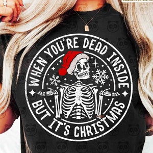 May include: Black t-shirt with a circular graphic featuring a skeleton wearing a Santa hat. The text reads "WHEN YOU'RE DEAD INSIDE BUT IT'S CHRISTMAS". The design includes snowflakes and stars.
