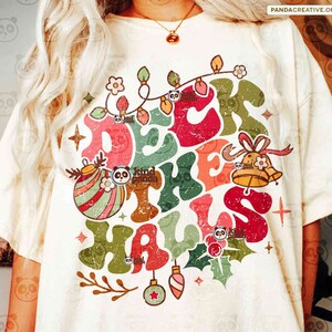 May include: A cream-coloured t-shirt with a vintage Christmas design. The design includes the phrase "Deck the Halls" in colourful, distressed lettering, festive ornaments, lights, and holly. The shirt also has the text "DIGITAL DOWNLOAD PNG DISTRESSED & SOLID PNGS".