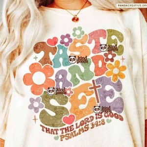 May include: A white t-shirt featuring a colourful graphic design that reads "Taste and See" with a cross and flowers. The graphic also includes the text "That the Lord is good, Psalms 34:8".