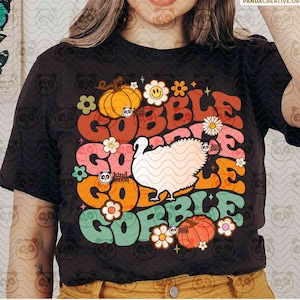 May include: A black short-sleeve t-shirt featuring a graphic print with the repeating text GOBBLE GOBBLE GOBBLE GOBBLE in orange, red, pink, and green. The design includes a white turkey illustration, orange pumpkins, and various colorful flowers.
