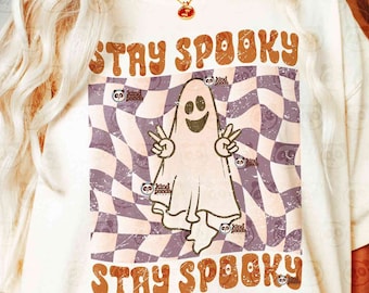 May include: A cream-colored t-shirt featuring a graphic of a ghost making a peace sign. The ghost is centered over a wavy, checkered pattern in shades of purple and white. The text "STAY SPOOKY" appears in a retro, orange-brown font above and below.
