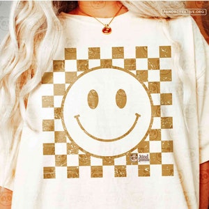 Peut inclure: Un t-shirt blanc avec un motif à carreaux dorés et un grand smiley doré au centre. Le texte "kind panda studio" est dans le coin inférieur droit.