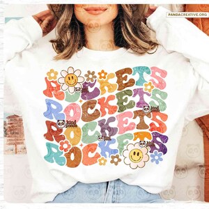 May include: A white sweatshirt with a colorful graphic design featuring the word "ROCKETS" in a retro 70s style font. The design includes flowers, smiley faces, and a panda logo.