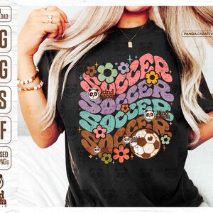 Op de afbeelding: Een zwart T-shirt met een kleurrijk grafisch ontwerp van het woord "soccer" in een retro-stijl met bloemen en pandaberen. De grafiek bevat ook een voetbal.