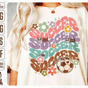 Op de afbeelding: Een wit T-shirt met een distressed grafisch ontwerp van het woord "soccer" in een regenboog van kleuren. Het ontwerp bevat bloemen en een voetbal met een hart erop. De tekst "kind panda studio" staat in het ontwerp.