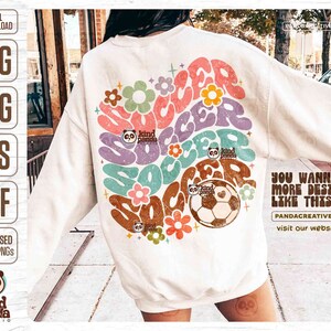 Op de afbeelding: Een wit sweatshirt met een retro-grafisch ontwerp met het woord "soccer" in kleurrijke, golvende letters. Het ontwerp bevat bloemen en een voetbal met een pandabeerlogo. De tekst "You wanna more design like this?" is gedrukt in de rechterbenedenhoek van het sweatshirt.