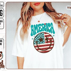 May include: A white t-shirt with a blue and red smiley face design. The smiley face is made up of the American flag and the text "AMERICA" is written in a retro font above the smiley face.