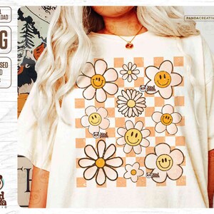 K&ouml;nnte beinhalten: Ein wei&szlig;es T-Shirt mit einem karierten Muster und einem Design aus mehreren l&auml;chelnden G&auml;nsebl&uuml;mchen in Rosa und Gelb. Der Text "kind panda" ist auf dem Design sichtbar.